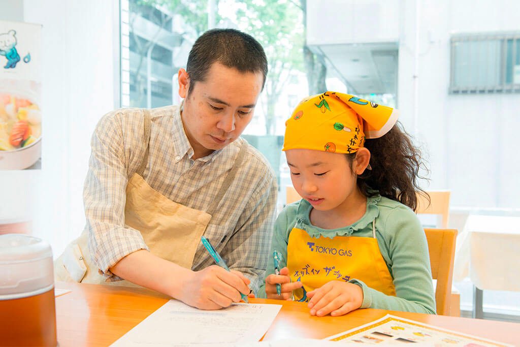 調理のレクチャーをメモをしながら真剣に聞／東京ガス キッズインザキッチン 親子クラス